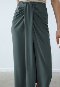 Green jersey maxi skirt with draped detailing and side slit, featuring a smooth, soft texture and gathered fabric at the waist.