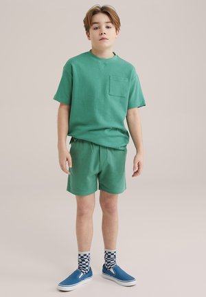T-shirt en coton vert avec poche, associé à un short assorti. Présente des chaussures bleues à enfiler et des chaussettes à carreaux, se tenant devant un fond neutre.
