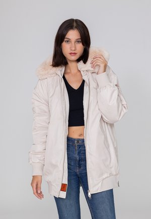 Veste bomber beige pâle avec un col en fausse fourrure, des poignets côtelés, une fermeture éclair et un patch étiqueté orange à l'ourlet. Le mannequin pose avec une main levée.