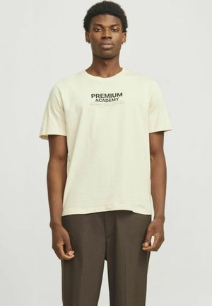 Cremefarvet t-shirt med korte ærmer, hvor "PREMIUM ACADEMY" er trykt i sort. Rund halsudskæring, lavet af bomuld, sat sammen med brune bukser.