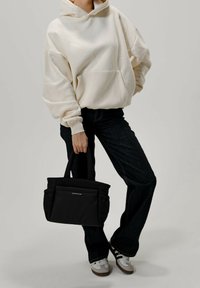 Svart tygkasse med två övre handtag, mjukt material, platt design, framfickor och en minimal textlogotyp. Bärs med en cremefärgad hoodie och mörka jeans.