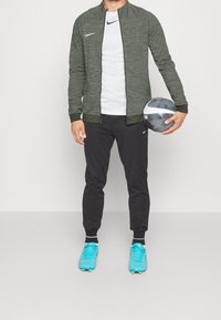 Groene jas met rits, witte blouse, zwarte broek en blauwe voetbalschoenen; houdt een zwart-wit voetbalsbal vast, relaxte fit.