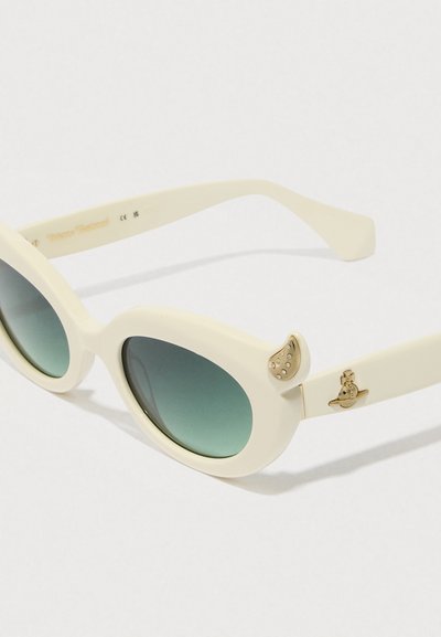 Óculos de sol de cor creme com lentes ovais, tonalidade verde degradê, detalhes decorativos em dourado e um design único de orelhas de gato nas laterais.
