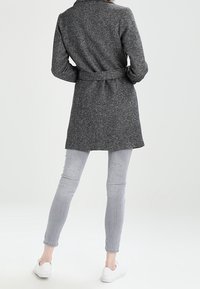 Grijze wolmix jas met een riem, voorzien van een kraag en een gestructureerd patroon, gedragen over lichtgrijze skinny jeans en witte sneakers.