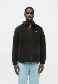 Barna kordbársony gombos ing gallérral, a mellkason kis zöld Lacoste logóval. Világoskék farmernadrág fölött viselve.