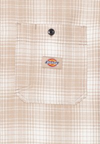 Kavos spalvos languota marškiniai su kišenėmis, kurių viršuje yra juodas mygtukas ir stačiakampis Dickies logotipo lipdukas.