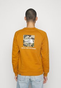 Sudadera naranja de algodón, con un logotipo de camuflaje cuadrado en la parte posterior y texto blanco, y acabados de canalé en el cuello y los puños.