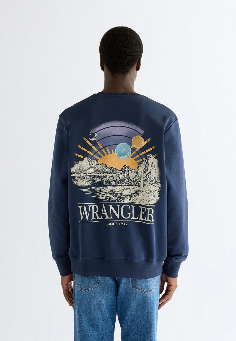 Personne portant un sweat-shirt bleu marine avec paysage désertique, planètes, rayons de soleil et texte "WRANGLER Since 1947" dans le dos, debout, dos tourné.
