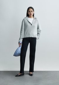 Woman standing against white wall, wearing gray knit sweater, black pants, black flats, holding soft blue handbag in left hand.