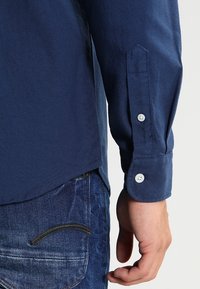 Höger arm och hand på en person som bär en långärmad marinblå skjorta med knäppt manschett och blå denimjeans.
