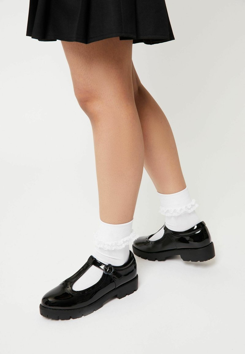 Fekete lakkbőr Mary Jane cipők T-pánttal, fehér fodros bok sockszal párosítva. A cipők alacsony, vastag sarkúak és kerek orrúak.