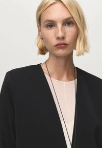 Blazer nero con scollatura a V sopra un top chiaro, caratterizzato da una texture liscia e un'accentuazione con collana nera. I capelli corti e biondi sono visibili.