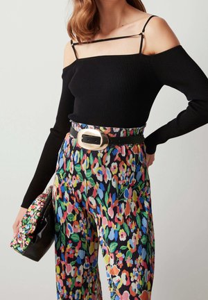 Woman wearing a black off-shoulder top with thin straps, multicolored floral pleated pants, wide black belt with large gold buckle, and matching clutch.