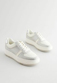 Weiße Sneakers mit silbernen Mesh-Einsätzen und perforierter Zehenpartie. Flache Schnürsenkel, gepolsterte Sohle und minimale silberne Akzente auf der Rückseite.