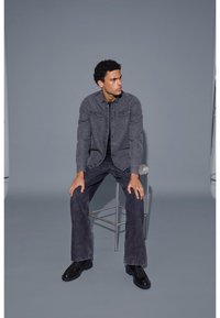Grå denimjacka över en svart skjorta, kombinerat med mörkgrå utsvängda jeans och svarta skor. Modellen sitter på en pall mot en neutral bakgrund.