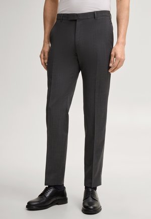 Mann trägt eng anliegende dunkelgraue Anzughose, schwarze Lederschuhe und ein weißes, in die Hose gestecktes Hemd vor neutralem Hintergrund.