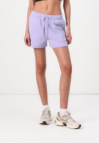 Shorts en coton lavande avec une taille élastique et un cordon de serrage. Présente un petit logo Nike blanc sur le devant. Portés avec des baskets beiges.