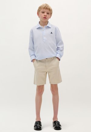 Camisa de manga larga azul claro con textura y cuello, pantalones cortos beige y mocasines negros. Detalle de botones notable en la tapeta de la camisa.
