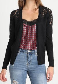 Kobieta ubrana w czarny koronkowy kardigan na czerwono-czarnym, kraciastym topie z koronkowym wykończeniem oraz niebieskie, postrzępione jeansy.