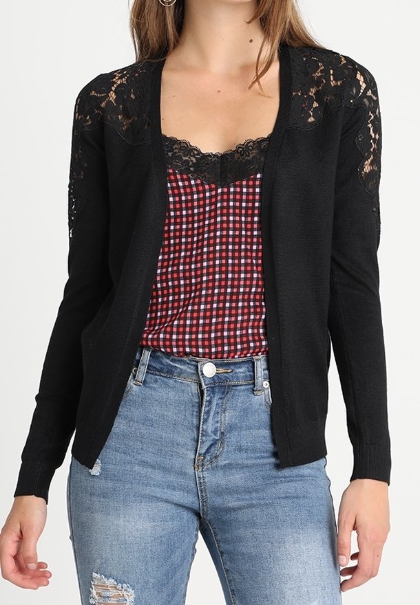 Kobieta ubrana w czarny koronkowy kardigan na czerwono-czarnym, kraciastym topie z koronkowym wykończeniem oraz niebieskie, postrzępione jeansy.
