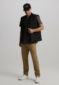 Czarna pikowana kamizelka na szarej koszulce, khaki spodnie i białe trampki; na głowie czapka w kolorze czarnym z logo. Swobodny strój, neutralna paleta kolorów.