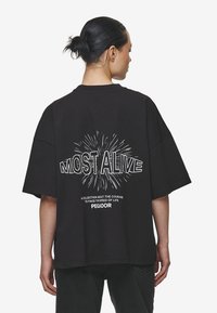 Zwarte oversized t-shirt met een ronde hals en korte mouwen. Kenmerkt zich door de witte opdruk "MOST ALIVE" en een explosie-ontwerp op de achterkant.