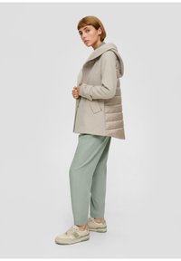 Femme portant une veste à capuche beige avec un dos matelassé, un pantalon vert clair et des baskets beiges, adoptant une pose décontractée sur un fond gris.