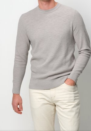 Mann trägt hellgrauen, strukturierten Langarmpullover und cremefarbene Hose, eine Hand in der Tasche, vor einfachem weißem Hintergrund.