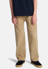 Enfant portant un pantalon en velours côtelé beige, des chaussures noires avec semelles en gomme, et une chemise foncée, debout les mains détendues le long du corps.