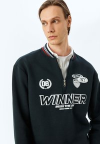 Sudadera con cremallera negra con el texto blanco "WINNER", cuello a rayas rojas y blancas, y parches que incluyen "NY" y el número "08". Tejido suave.