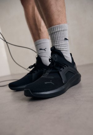 Sapatos desportivos pretos com parte superior em malha, sola de borracha e atacadores. Usados com meias cinzas caneladas com um pequeno logotipo. O fundo é neutro.