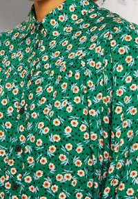 Groene bloemenshirt met kleine oranje bloemetjes en zwarte stippen, met een knoopsluiting aan de voorkant en een gestructureerde kraag. Zacht stof met plooien.