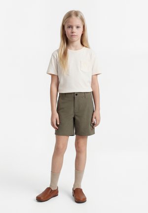 Ξανθό παιδί που στέκεται με το πρόσωπο προς τα εμπρός, φορώντας μπεζ κοντομάνικο πουκάμισο, χακί σορτσάκι, μπεζ κάλτσες και καφέ παπούτσια χωρίς κορδόνια σε λευκό φόντο.