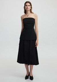 Schwarzes trägerloses Kleid mit tailliertem Oberteil und Peplum-Taille, das in einen voluminösen Rock übergeht. Glatte Textur, kombiniert mit schwarzen spitzen Absätzen.