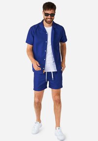 Camicia a maniche corte blu con bottoni, t-shirt bianca sotto, pantaloni corti blu, sneakers bianche. Tessuto liscio, design casual, maniche corte e pantaloni corti arrotolati.