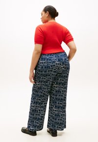 Κόκκινο, κοντομάνικο πουλόβερ με υφή, συνδυασμένο με μπλε σκούρο φαρδιά παντελόνια με γεωμετρικό λευκό μοτίβο. Μαύρα παπούτσια ολοκληρώνουν την εμφάνιση.