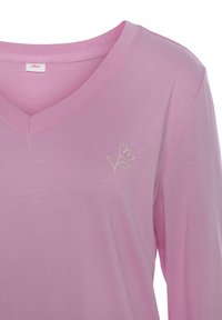 Rosa V-Ausschnitt-T-Shirt aus weichem Stoff, mit einer kleinen weißen Blumenstickerei auf der linken Seite. Einfaches Design mit langen Ärmeln.
