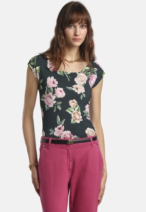 Donna con capelli castani ondulati che indossa una maglia nera a fiori, pantaloni rosa con cintura nera e gioielli dorati su uno sfondo chiaro uniforme.