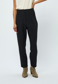 Pantaloni neri a vita alta con pieghe e tasche laterali, caratterizzati da una texture liscia e un design a gamba affusolata. Abbinati a stivali alla caviglia color taupe.