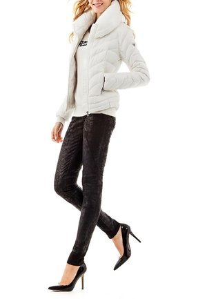 Nainen, joka pukeutunut valkoiseen tikattuun takkiin, mustiin teksturoituihin leggingseihin ja mustiin korkokenkiin, poseeraa siten, että yksi jalka on ristissä toisen edessä.