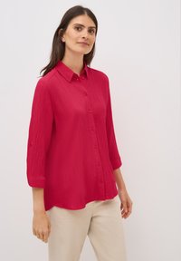 Camisa roja de botones con una tela texturizada, mangas enrolladas y dobladillo curvado. Llevada sobre pantalones de color claro.