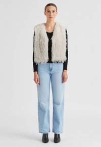Faux-Fur-Weste in Off-White mit einer wolligen Textur, getragen über einem schwarzen Langarmshirt, kombiniert mit hellblauen Jeans im geraden Schnitt und schwarzen Stiefeln.