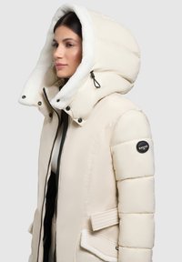 Crème kleurige pufferjack met een grote, pluche capuchon gevoerd met witte fleece. Voorzien van drukknoopsluitingen, ritsen en een logo patch op de mouw.