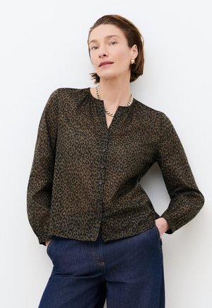 Vrouw met een donker groen blouse met luipaardprint en knopen, een gouden ketting, gouden oorringen en donkerblauwe spijkerbroek, met handen in de zakken.