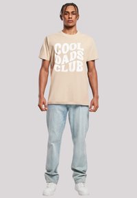 Camiseta de algodón beige con "COOL DADS CLUB" impreso en blanco, combinada con jeans de mezclilla de lavado claro y zapatillas blancas.