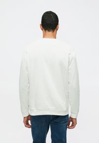 Felpa bianca con maniche lunghe, polsini e orlo a coste. La vista posteriore mostra una texture semplice e liscia e una vestibilità comoda. Abbinata a jeans blu.