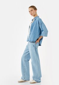 Camisa de denim de corte oversize en azul claro con cierre de botones, mangas de tres cuartos, combinada con pantalones anchos en azul claro y zapatos beige.