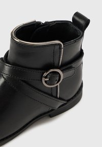 Botte noire en cuir au niveau de la cheville avec une boucle argentée ronde, des coutures décoratives et une tige texturée. Elle possède un petit tirant à l'arrière.