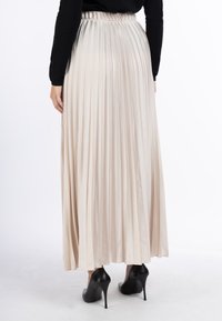 Plisseret maxiskørt i lys beige satin med elastisk talje. Båret med sorte højhælede sko. Glat tekstur og flydende design.
