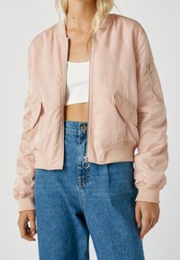 Femme blonde portant une veste bomber rose clair sur un crop top blanc côtelé et un jean taille haute bleu, sur un fond uni.